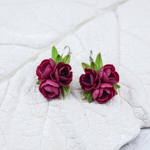May include: A pair of burgundy flower earrings with green leaves. The earrings are made of polymer clay and have a silver hook.