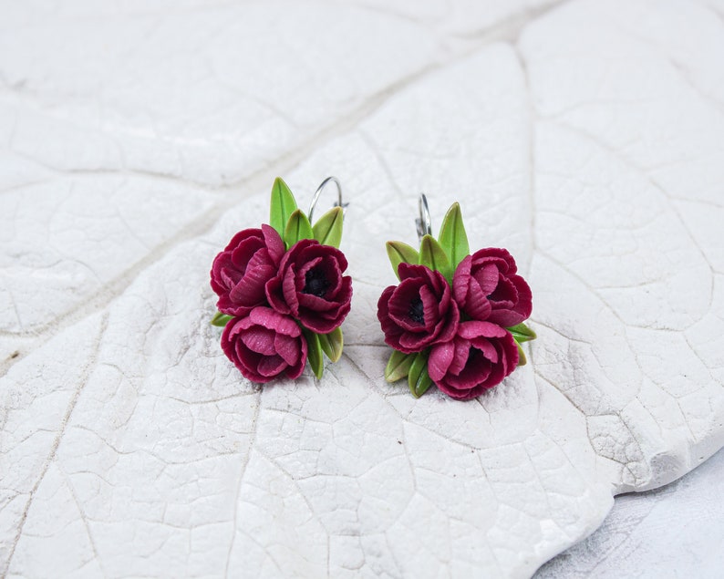 May include: A pair of burgundy flower earrings with green leaves. The earrings are made of polymer clay and have a silver hook.