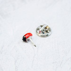 May include: A small lapel pin shaped like a ladybug with a glossy red and black body. The pin features a silver-toned metal post and a separate circular metal clutch backing, designed to secure the accessory through fabric or other materials.