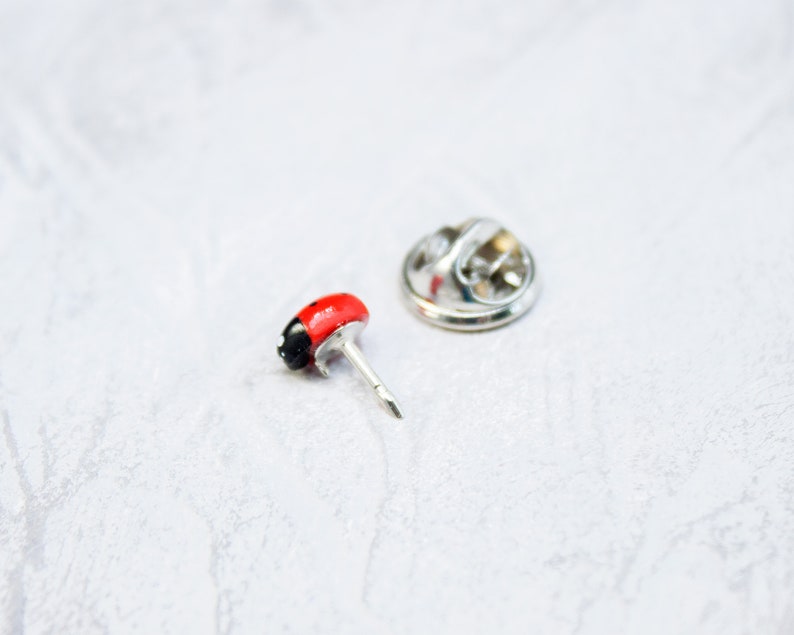 May include: A small lapel pin shaped like a ladybug with a glossy red and black body. The pin features a silver-toned metal post and a separate circular metal clutch backing, designed to secure the accessory through fabric or other materials.