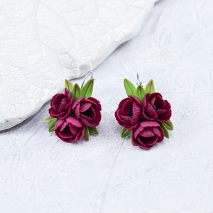 May include: A pair of burgundy flower earrings with green leaves. The earrings are made of polymer clay and have a delicate, feminine look.