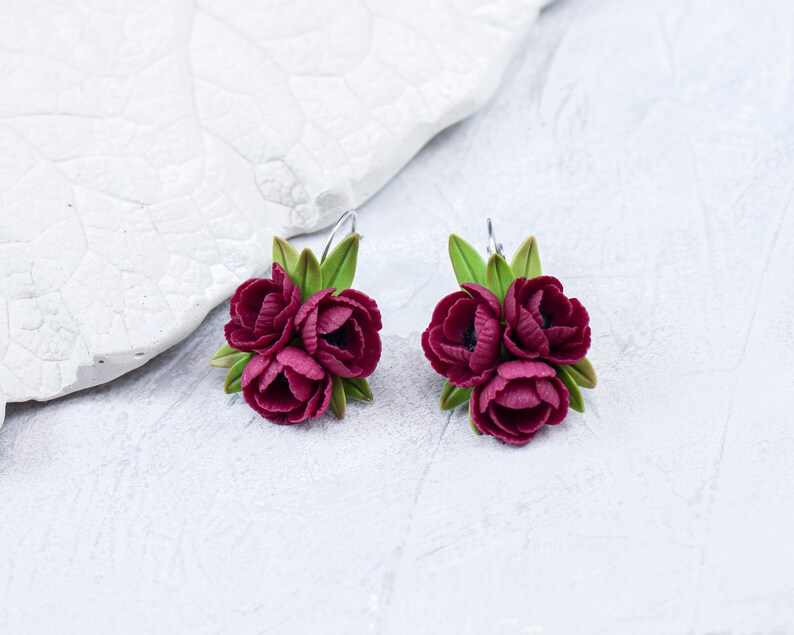 May include: A pair of burgundy flower earrings with green leaves. The earrings are made of polymer clay and have a delicate, feminine look.