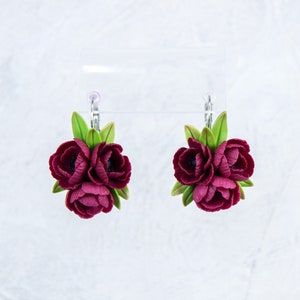May include: A pair of earrings featuring three burgundy polymer clay flowers with green leaves. The earrings are attached to silver hooks.
