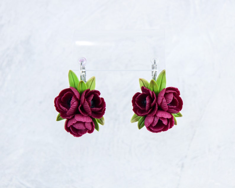 May include: A pair of earrings featuring three burgundy polymer clay flowers with green leaves. The earrings are attached to silver hooks.