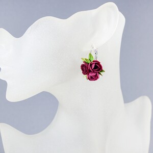 May include: A pair of earrings featuring a cluster of three burgundy-colored fabric flowers with green leaves. The earrings are hanging from silver hooks.