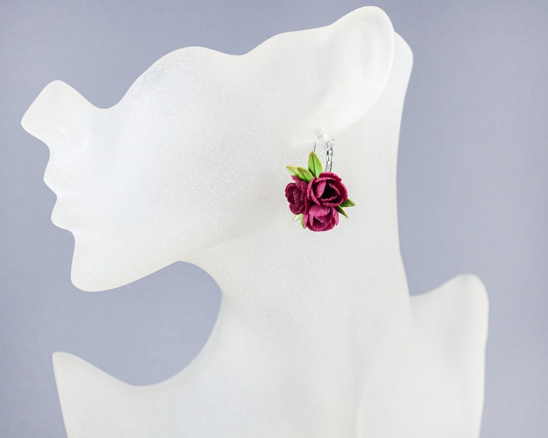 May include: A pair of earrings featuring a cluster of three burgundy-colored fabric flowers with green leaves. The earrings are hanging from silver hooks.