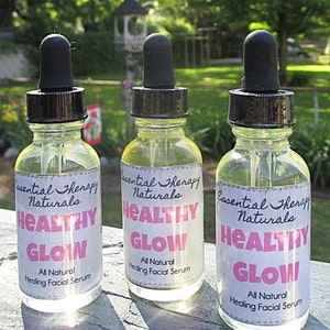 Puede incluir: Tres botellas de vidrio transparente con cuentagotas negros, etiquetadas como "Essential Therapy Naturals Healthy Glow All Natural Healing Facial Serum". Contienen un l&iacute;quido amarillo, sobre un fondo verde borroso.