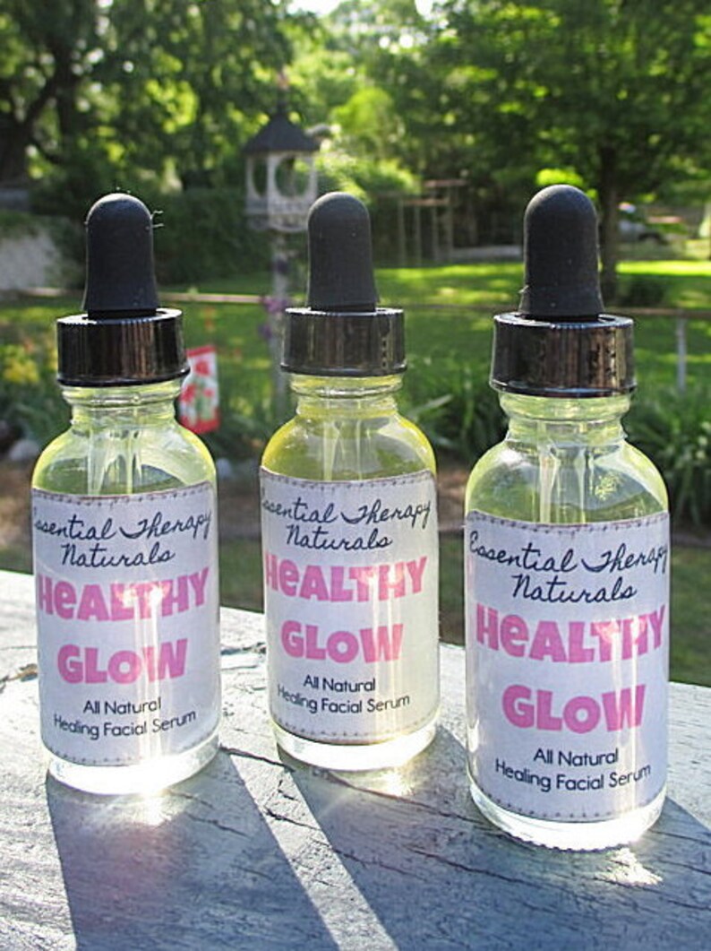 Puede incluir: Tres botellas de vidrio transparente con cuentagotas negros, etiquetadas como "Essential Therapy Naturals Healthy Glow All Natural Healing Facial Serum". Contienen un l&iacute;quido amarillo, sobre un fondo verde borroso.
