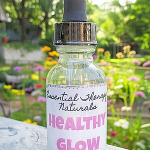 Puede incluir: Una botella de vidrio transparente con un cuentagotas negro, etiquetada como "Essential Therapy Naturals Healthy Glow All Natural Healing Facial Serum". La botella se encuentra sobre un fondo borroso de vegetaci&oacute;n y flores.