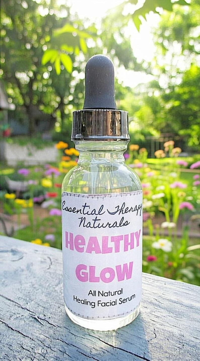 Puede incluir: Una botella de vidrio transparente con un cuentagotas negro, etiquetada como "Essential Therapy Naturals Healthy Glow All Natural Healing Facial Serum". La botella se encuentra sobre un fondo borroso de vegetaci&oacute;n y flores.