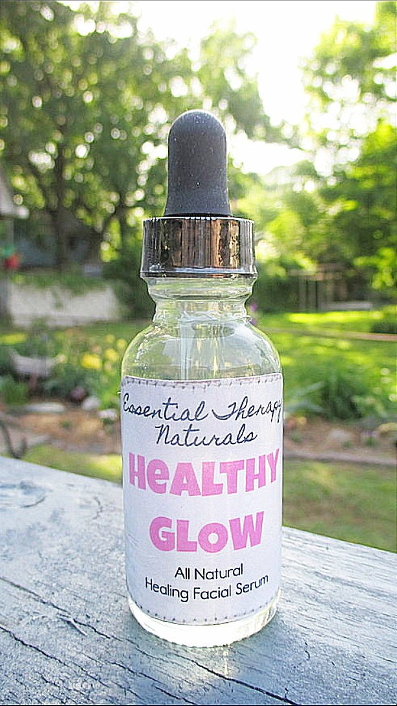 Puede incluir: Una botella de vidrio transparente con un cuentagotas negro y un collar de metal plateado. La botella tiene una etiqueta blanca con las palabras "Essential Therapy Naturals" y "HEALTHY GLOW" en rosa. La etiqueta tambi&eacute;n dice "All Natural Healing Facial Serum".