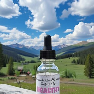 Puede incluir: Una botella de vidrio transparente con un cuentagotas negro, etiquetada como "Essential Therapy Naturals HEALTHY GLOW All Natural Healing Facial Serum". El fondo presenta una vista panor&aacute;mica de monta&ntilde;as, &aacute;rboles y un cielo azul con nubes.