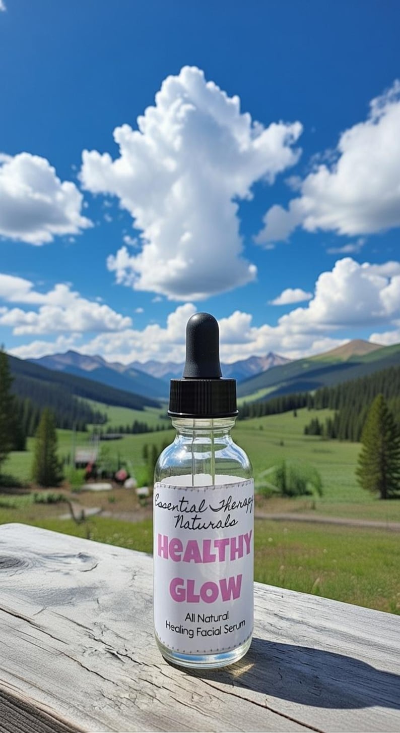 Puede incluir: Una botella de vidrio transparente con un cuentagotas negro, etiquetada como "Essential Therapy Naturals HEALTHY GLOW All Natural Healing Facial Serum". El fondo presenta una vista panor&aacute;mica de monta&ntilde;as, &aacute;rboles y un cielo azul con nubes.