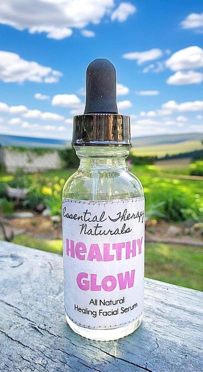 Puede incluir: Una botella de vidrio transparente con un cuentagotas negro, etiquetada como "Essential Therapy Naturals". La etiqueta presenta las palabras "HEALTHY GLOW" en rosa y "All Natural Healing Facial Serum". El fondo muestra un cielo azul y follaje verde.
