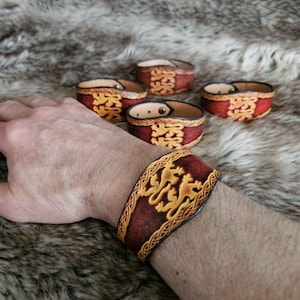 Op de afbeelding: Een brede bruine leren manchetarmband met een centraal reliëfontwerp van twee klimmende leeuwen omringd door een decoratieve gevlochten rand. De armband is voorzien van een metalen drukknoopsluiting en heeft donker afgewerkte randen.