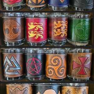 Puede incluir: Una colección de vasos de cristal envueltos en fundas de cuero repujado con diversos diseños, incluyendo una triqueta roja, el símbolo de las Reliquias de la Muerte, un logo de Superman, un emblema de Batman, árboles, leones, un valknut, un pentagrama, un trisquel, una cruz, un jabalí, una calavera y un ciervo.
