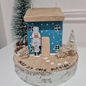 May include: A wooden house painted blue with white polka dots and a nutcracker figurine standing in the doorway. The house is sitting on a wooden base with the words "Baby it's cold outside." painted on it.