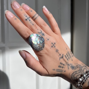 May include: A large, oval-shaped ring featuring a polished abalone shell inlay. The shell displays a natural, iridescent surface with swirling patterns of deep blue, green, purple, and silver tones set within a smooth, rounded metal frame.