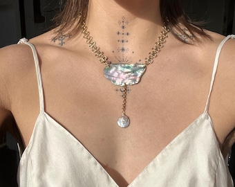 May include: A gold-toned chain necklace featuring a central iridescent abalone shell pendant with a cloud-like shape. A delicate gold chain hangs from the center of the shell, ending in a small, round, white coin-shaped pearl charm.