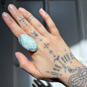 May include: A large oval ring featuring a light green and purple stone. The ring is set on a hand with multiple tattoos, including stars, crosses, and a crescent moon. The ring is the focal point of the image.