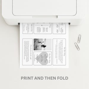May include: A printed wedding invitation emerges from a white printer. The invitation features text and a black and white photo. The text includes wedding details and the phrase "Our Love Story". Two silver paper clips are on the white surface.