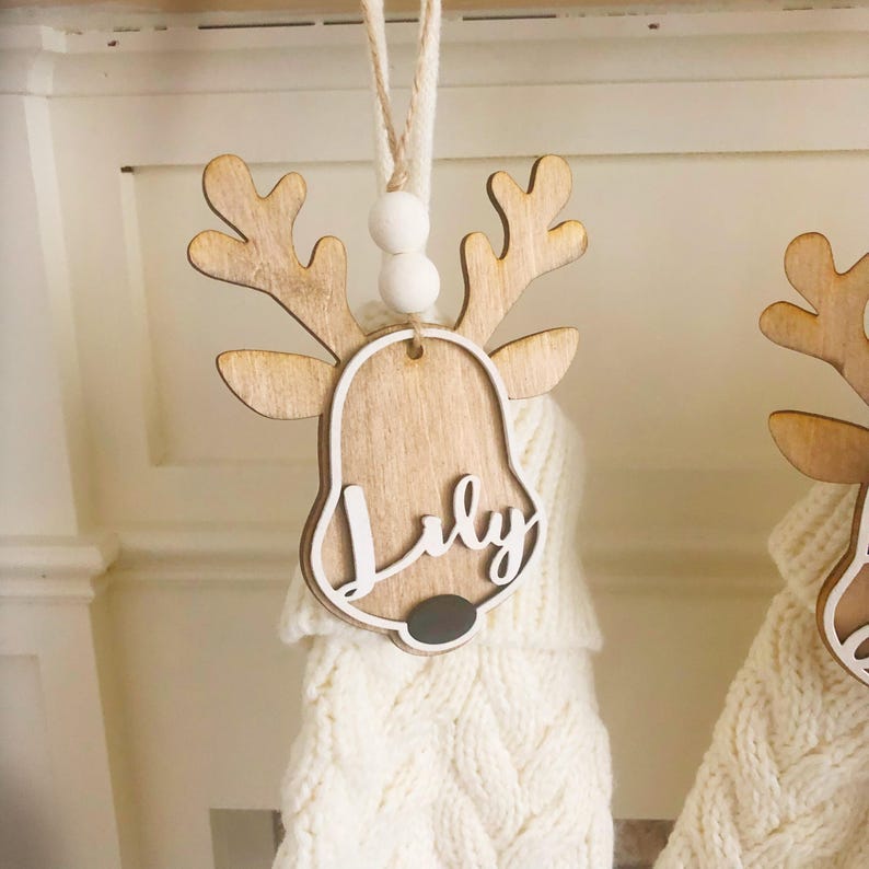 May include: A wooden reindeer ornament featuring a light brown finish, white trim, and a dark grey nose. The name Lily is written in white script across the centre. The ornament hangs from a twine string with two white wooden beads above the head.