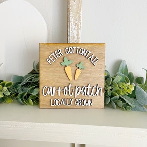 May include: Wooden sign with the text "Peter Cottontail carrot patch locally grown" in white lettering. Two small orange carrots with green tops are above the text.