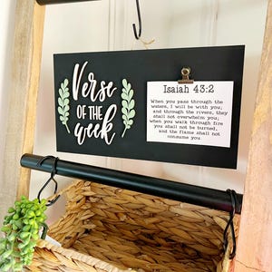 May include: A black sign with the words "Verse of the Week" in white script and decorative green leaves. A smaller white paper with text from Isaiah 43:2 is clipped to the sign. The sign hangs on a black bar above a woven basket.