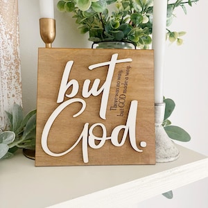 May include: A square wooden sign with a light brown finish featuring white raised lettering that reads but God. Smaller black text integrated into the design reads There was no way, but GOD made a way. The sign has a smooth, flat surface.