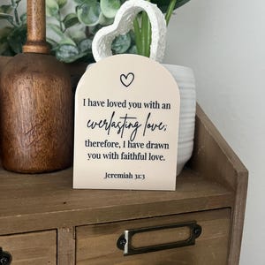 May include: A cream-coloured, arched plaque with a heart symbol and the text "I have loved you with an everlasting love; therefore, I have drawn you with faithful love. Jeremiah 31:3." The plaque has a heart-shaped handle.