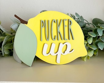 May include: A yellow lemon-shaped wooden sign with the text "pucker up" in green and white lettering.