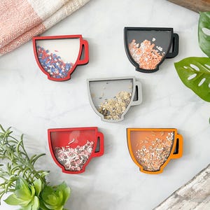 May include: Five wooden teacup decorations in various colours. Each cup has a different coloured frame: red, black, grey, red, and orange. The clear centres are filled with confetti in various colours and shapes, creating a playful, decorative effect.
