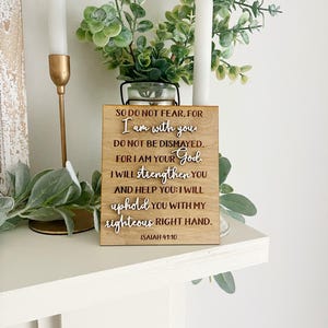 May include: A wooden sign with the text "SO DO NOT FEAR, FOR I am with you..." is displayed on a white surface. The sign is accompanied by greenery, candles, and a gold candle holder, creating a decorative arrangement.