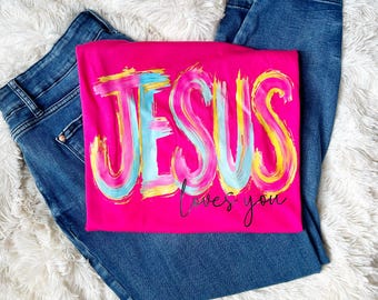 May include: A hot pink t-shirt with the word "JESUS" in a colorful, brushstroke design. Below the word "JESUS" is the phrase "loves you" in a handwritten font. The shirt is paired with blue denim jeans.