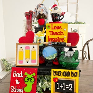 May include: A tiered display decorated with back to school themed items. The display features a gnome holding an apple, a coffee mug with the letters A+ on it, a sign that says "Love Teach Inspire Back 2 School", a sign with three pencils, a sign that says "Back to School" with a green worm wearing glasses, and a sign that says "Teacher Gonna Teach 1+1=2".