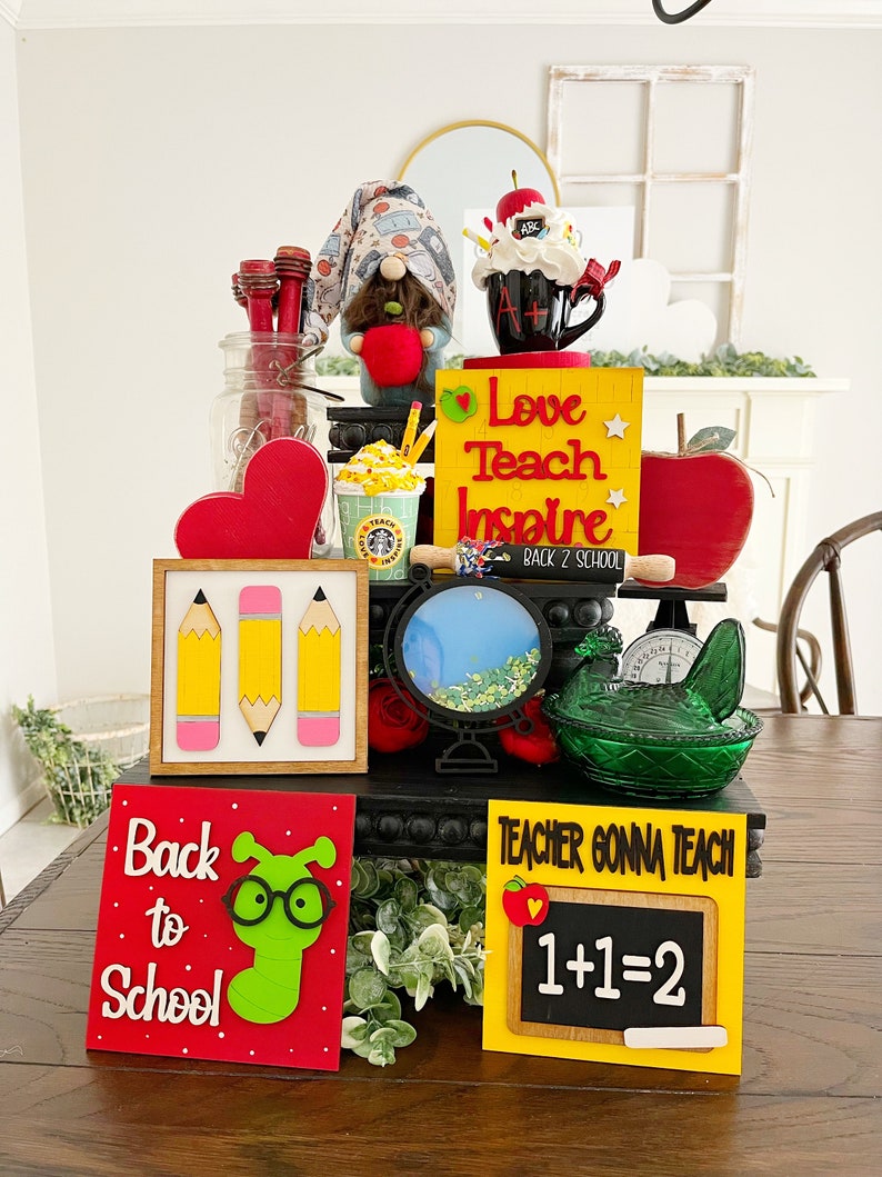 May include: A tiered display decorated with back to school themed items. The display features a gnome holding an apple, a coffee mug with the letters A+ on it, a sign that says "Love Teach Inspire Back 2 School", a sign with three pencils, a sign that says "Back to School" with a green worm wearing glasses, and a sign that says "Teacher Gonna Teach 1+1=2".