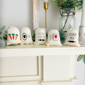 May include: Five small, off-white fabric bags with black and red graphics and text. The bags are decorated with images of carrots, an eye, a heart stamp, and text that reads "CHRISTMAS COOKIE MIX" and "COOKIE MIX". The bags are displayed on a white shelf.