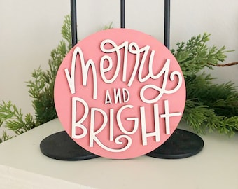 May include: Pink round wooden sign with white lettering that reads "merry and bright".