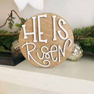 May include: A round wooden sign with the words "He is Risen" in white lettering. The sign is on a white shelf with green foliage and silver ornaments.