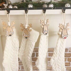 May include: Four cream-coloured cable knit stockings hanging from black hooks. Each stocking features a wooden reindeer ornament with white beads and a name tag reading Lily, Jace, Johnny, or Sara. The reindeer have white script lettering.