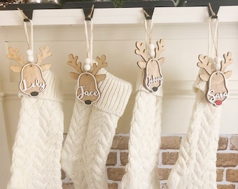 May include: Four cream-colored cable knit stockings hanging from black hooks. Each stocking features a wooden reindeer ornament with white beads and a name tag reading Lily, Jace, Johnny, or Sara. The reindeer have white script lettering.