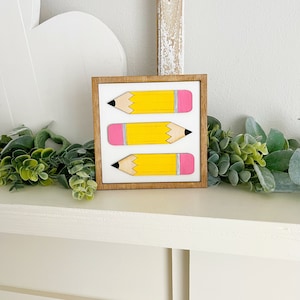 May include: A framed wooden sign with three yellow pencils with pink erasers and black lead tips. The pencils are arranged vertically in a row.