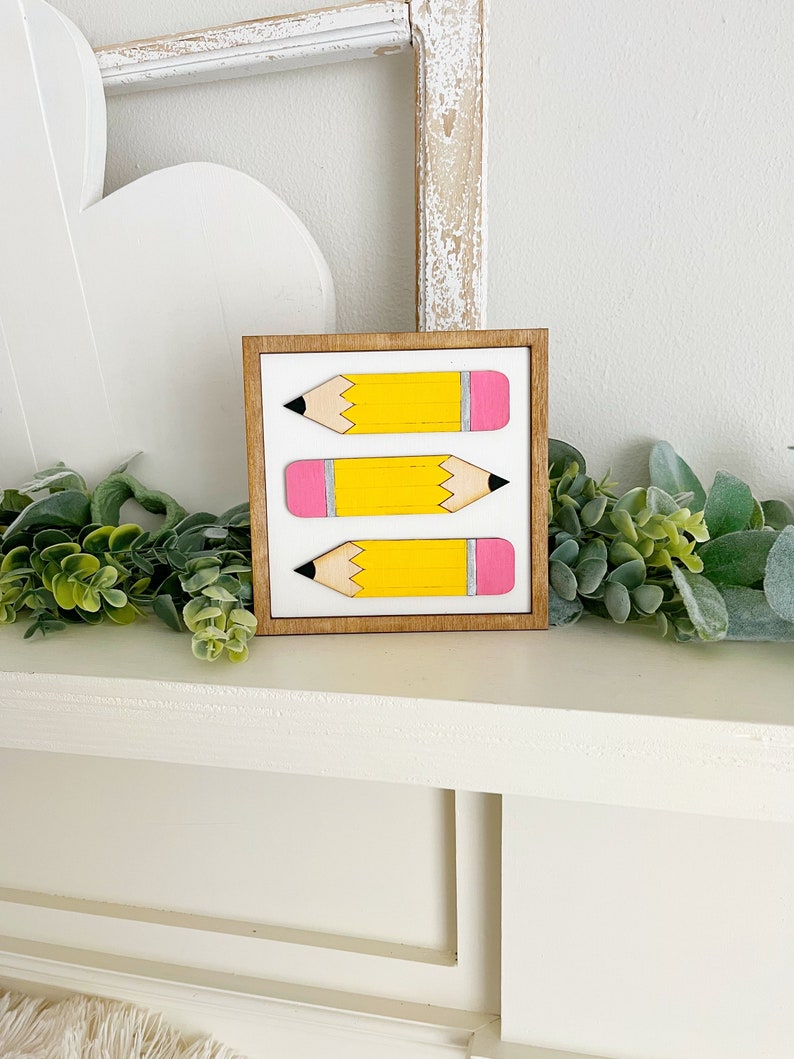 May include: A framed wooden sign with three yellow pencils with pink erasers and black lead tips. The pencils are arranged vertically in a row.