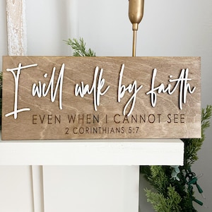May include: A rectangular wooden sign with a light brown stained finish featuring the white 3D lettering "I will walk by faith" above the engraved text "EVEN WHEN I CANNOT SEE" and "2 CORINTHIANS 5:7" in a clean, sans-serif font style.