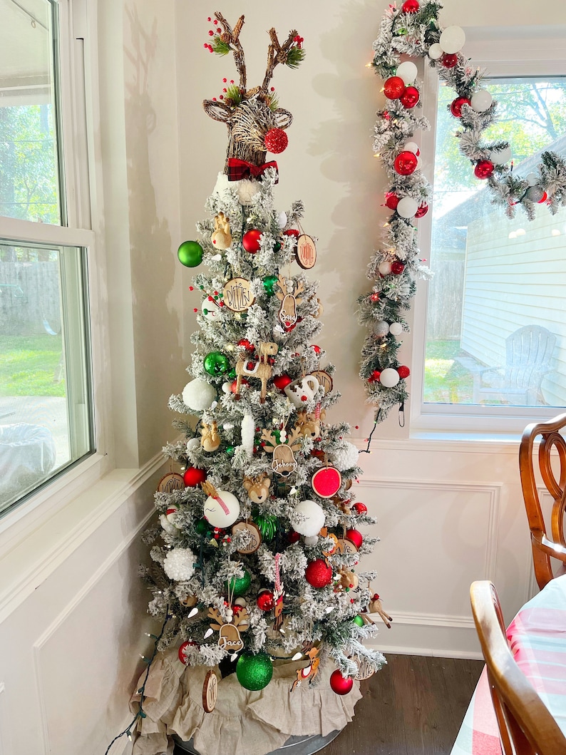May include: A flocked Christmas tree decorated with red and green baubles, wooden name tags, and various reindeer-themed accents. A large reindeer head topper with a red nose and a plaid bow sits at the peak, while a matching garland hangs nearby.