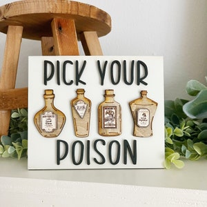 May include: White wooden sign with black text that reads "Pick Your Poison". Four wooden bottles with labels are attached to the sign. The labels read "High Quality Small Slime", "Elixir of Life", "Dragon's Breath", and "The Unforgiven".