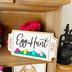 May include: Wooden sign with the text "Egg Hunt" in black script. The sign is shaped like a ticket with the words "Admit One" on the right side. There are five colorful Easter eggs on a green grass background.