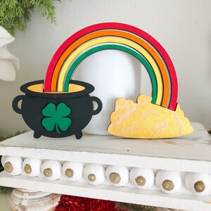 May include: A wooden rainbow with a pot of gold at the end. The pot is black with a green shamrock on the front. The rainbow is made of red, orange, yellow, green, and blue wood.