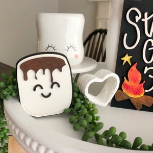 May include: A white ceramic mug with a smiling face and eyelashes, a white heart-shaped marshmallow, a wooden marshmallow with chocolate drizzle, and a wooden sign with the text "S'mores" and a painted campfire.