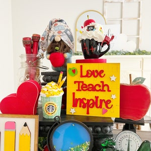 May include: A teacher-themed tabletop display with a gnome holding an apple, a coffee mug with an A+ on it, a yellow sign that says "Love Teach Inspire", a red apple, a globe, a green glass chicken, and a scale.